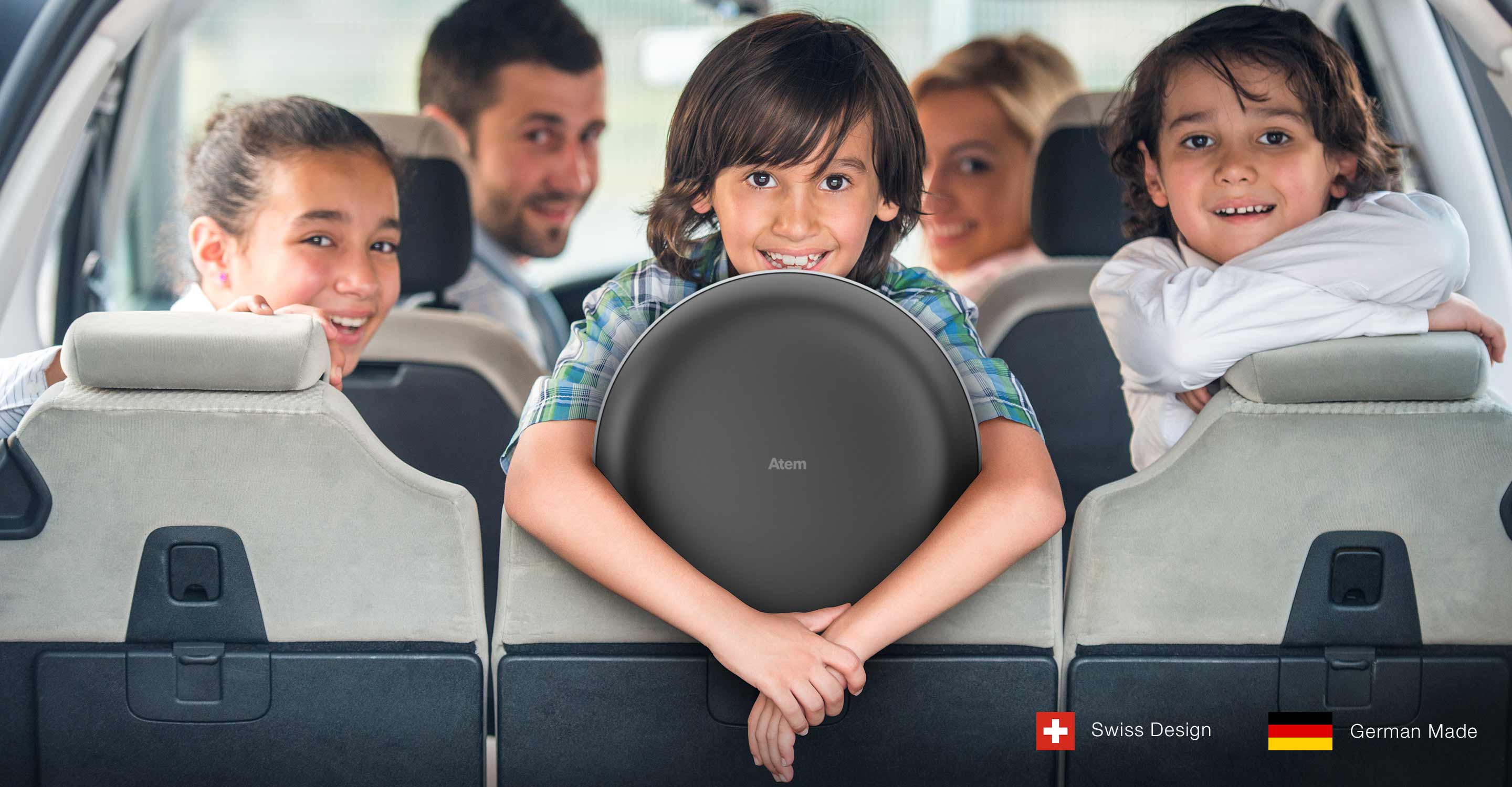 Family of five in car, seen from vantage point of back of the car, with all family members looking straight into the camera, smiling, with an Atem Car mounted on the backseat, embraced by a long-haired child.